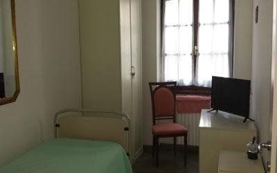 Una piccola camera da letto minimalista con un letto singolo con lenzuola verde chiaro, un armadio alto bianco, una sedia e un televisore.