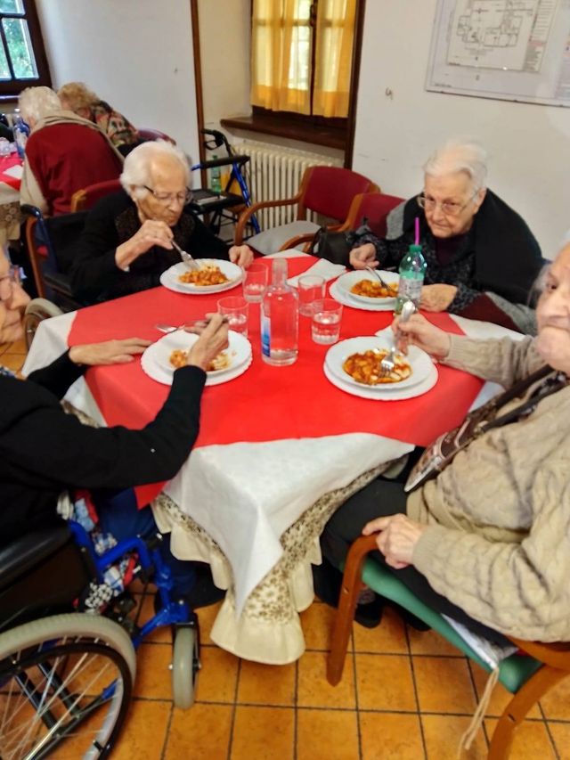 In una sala da pranzo ben illuminata, le persone pranzano insieme attorno a un tavolo quadrato coperto da una tovaglia rossa.