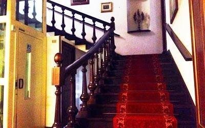 Una scala rivestita di moquette rossa con ringhiere in legno scuro all'interno di un edificio, caratterizzata da una nicchia a muro con una piccola scultura in metallo.