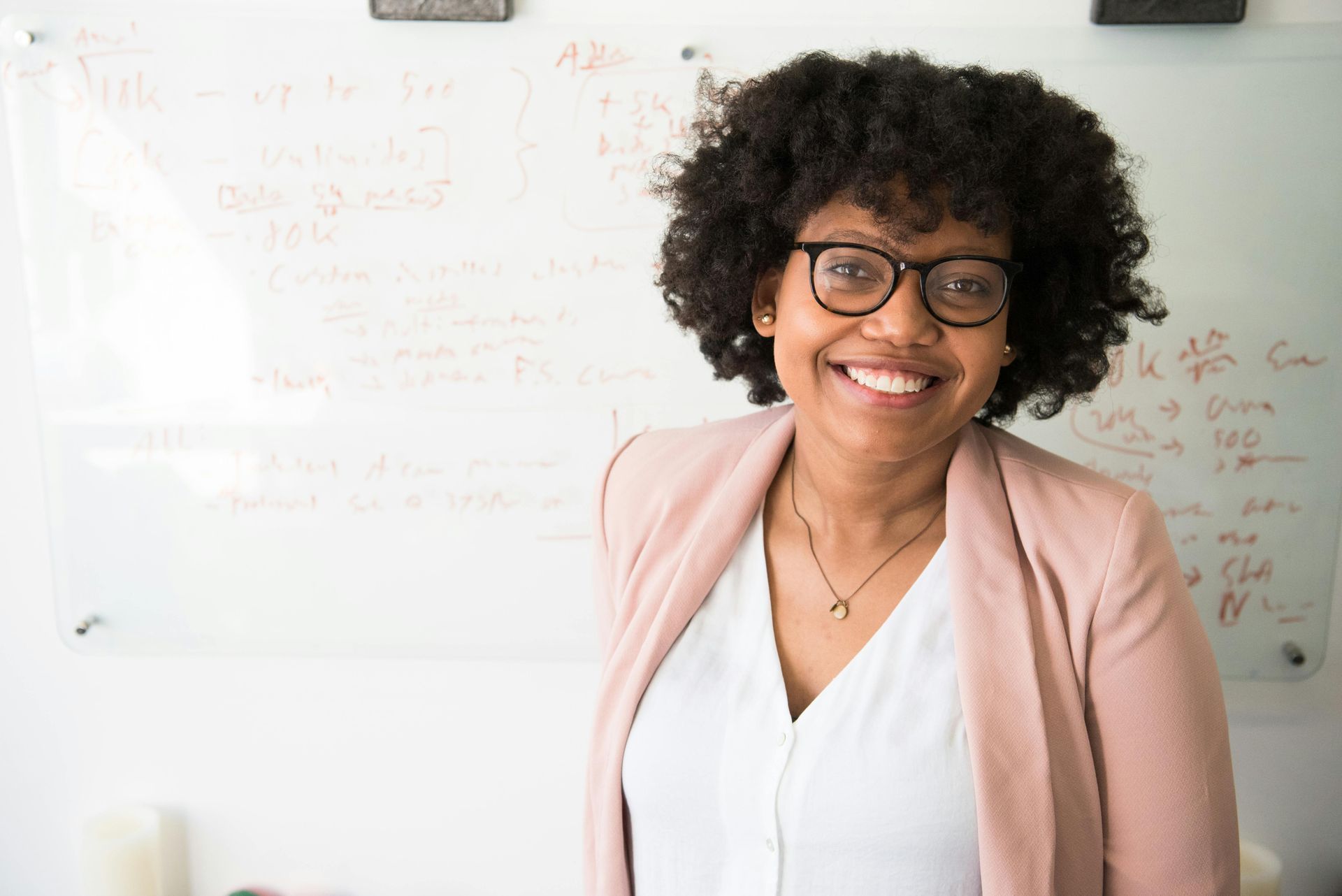 Een vrouw met een bril staat lachend voor een whiteboard.