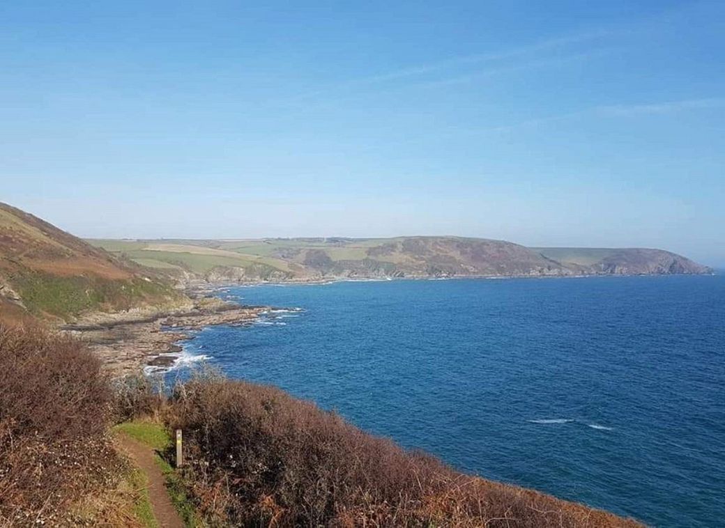 View from a clifftop path into a wide bay with blue water, blue sky and high cliffs beyond towards Polperro