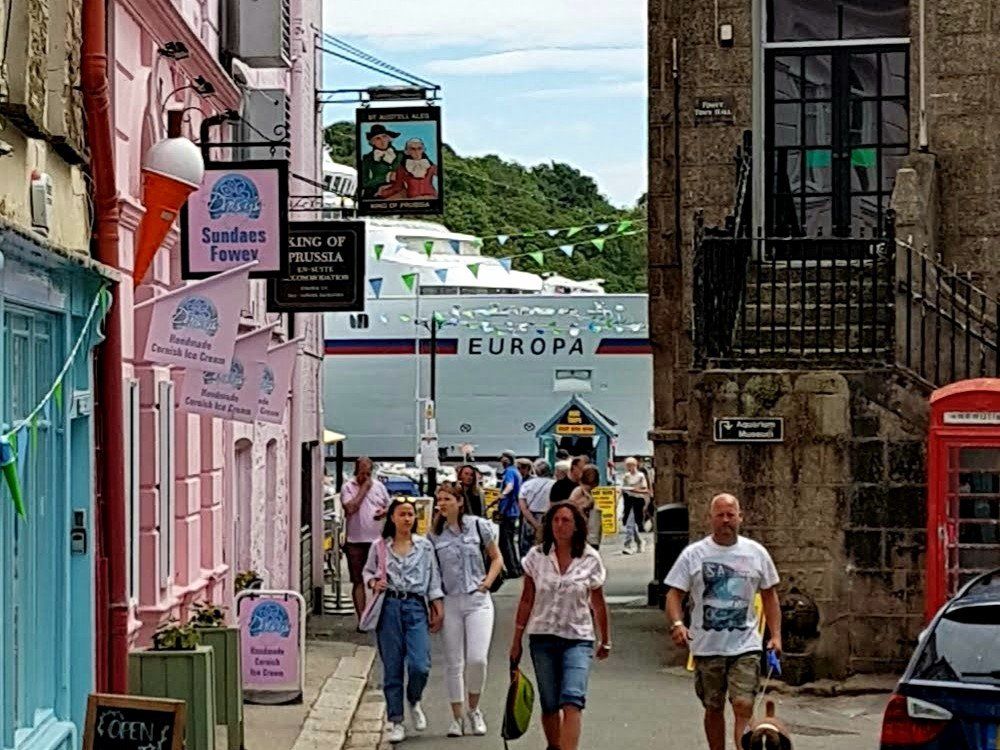 A busy street between ancient buildings looking towards a large cruiseship moored in the Fowey river
