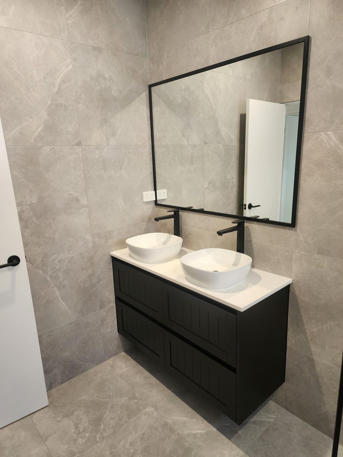 Modern bathroom vanity with double sinks, black cabinet, large mirror, and gray tiled walls.