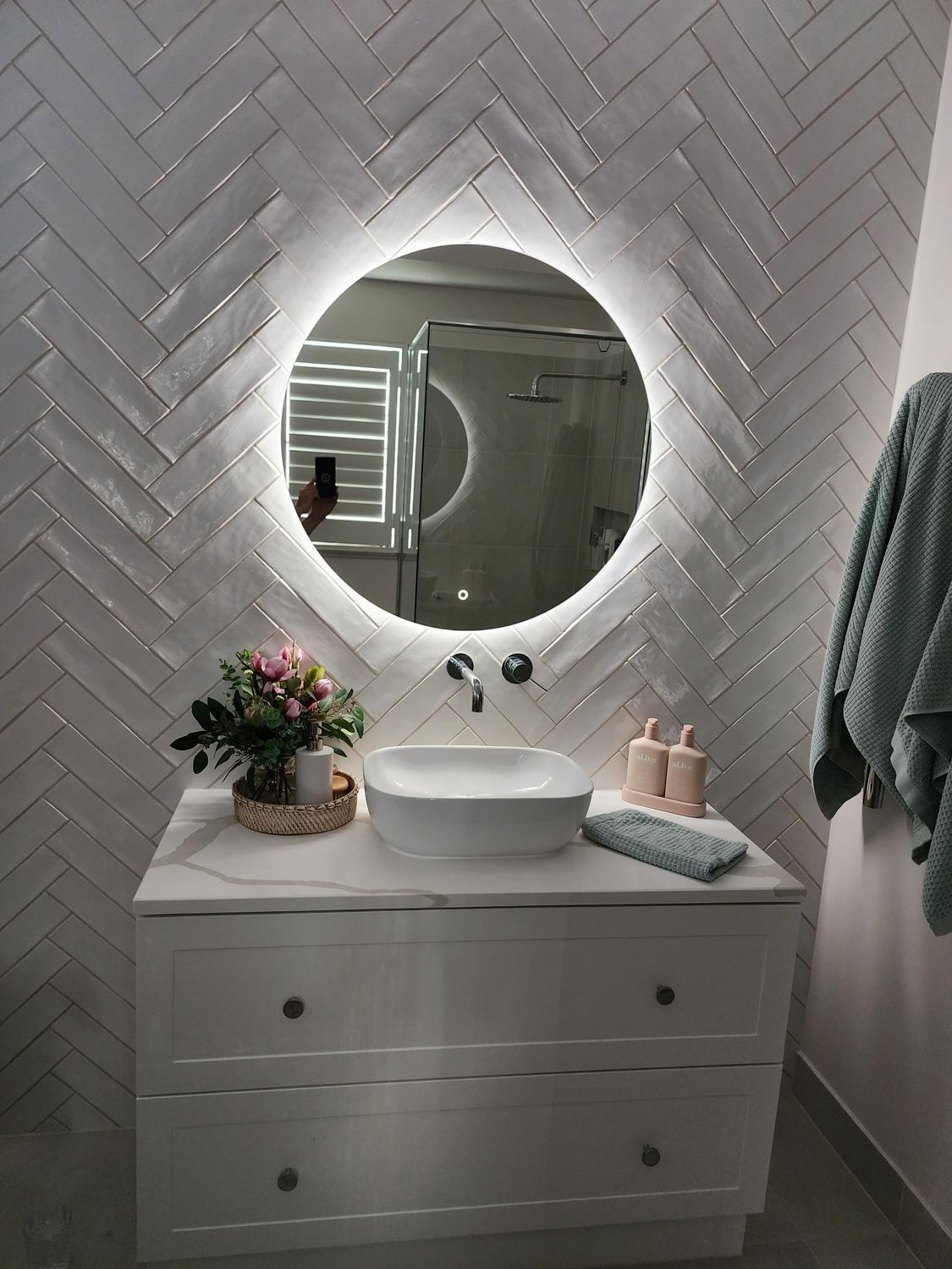 Bright bathroom vanity with round mirror, white sink, herringbone tile wall, and gray towels