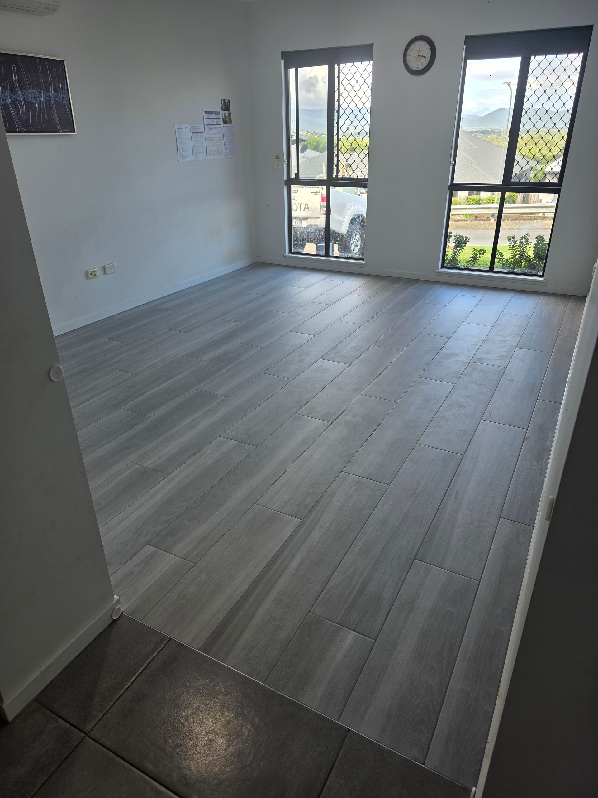 Empty bright room with light wood flooring, white walls, and large windows overlooking greenery.