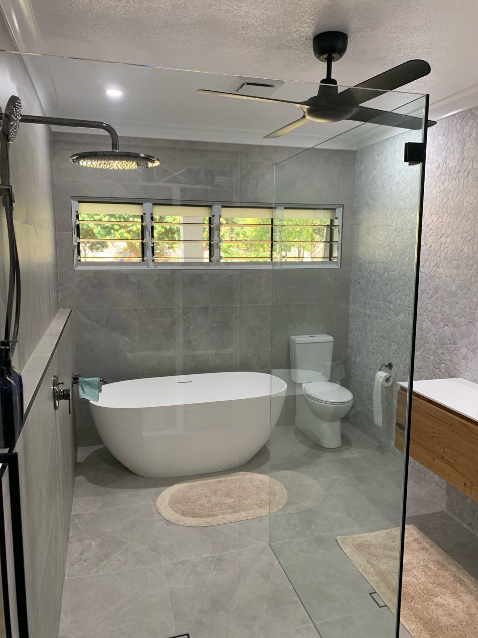 Modern gray bathroom with a freestanding tub, toilet, walk-in shower, window, and ceiling fan