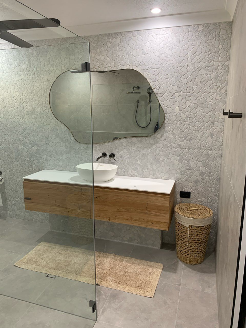 Modern bathroom with floating wood vanity, round mirror, glass shower screen, and wicker hamper
