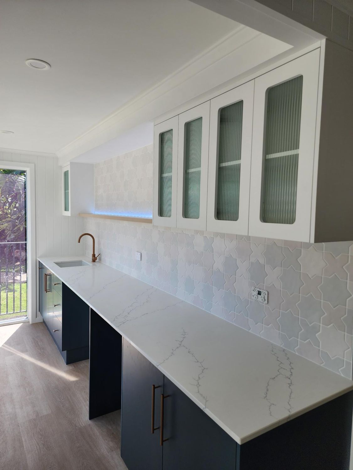 Modern kitchen with long white countertop, white cabinets, and light gray tiled backsplash by a windowed wall.