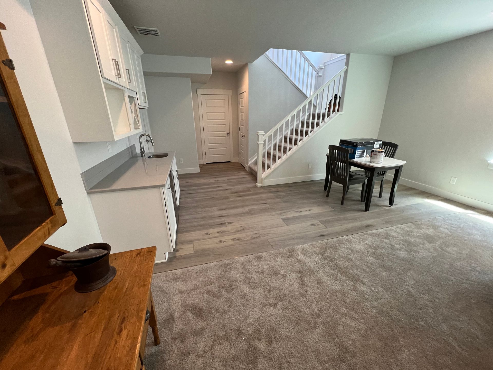A room featuring a white kitchen corner, wood-look flooring, a staircase with white railings, and a small dining set.