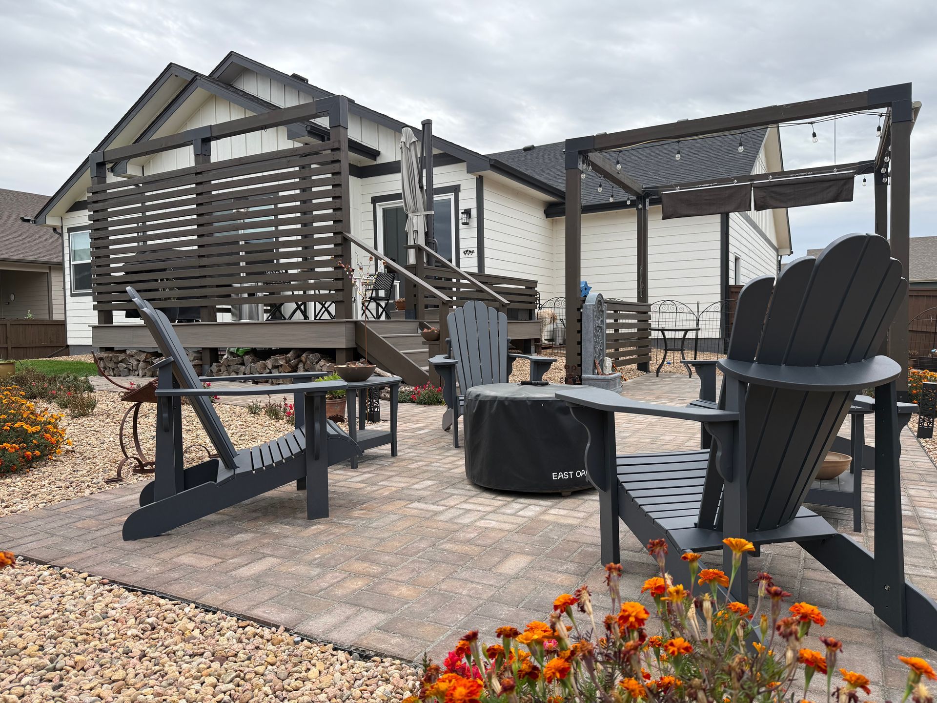 A patio with dark Adirondack chairs, a fire pit, and a wooden pergola set against the back of a light-colored home.