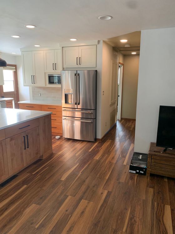 A kitchen with wood-toned cabinets and flooring, a stainless steel refrigerator, and a doorway leading to a hallway.