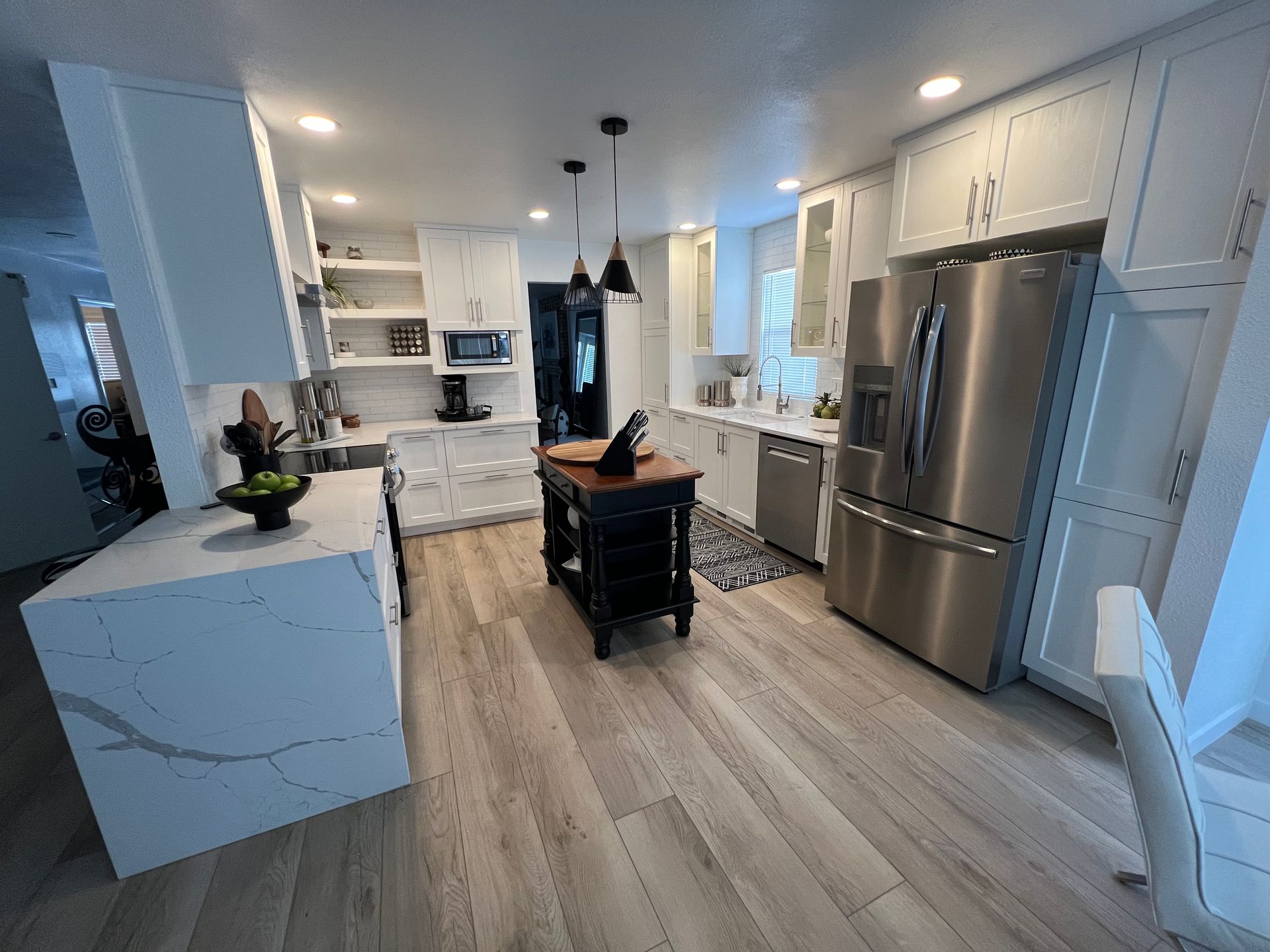 A modern white kitchen with a quartz island, dark wood center island, stainless steel refrigerator, and wood-look floors.