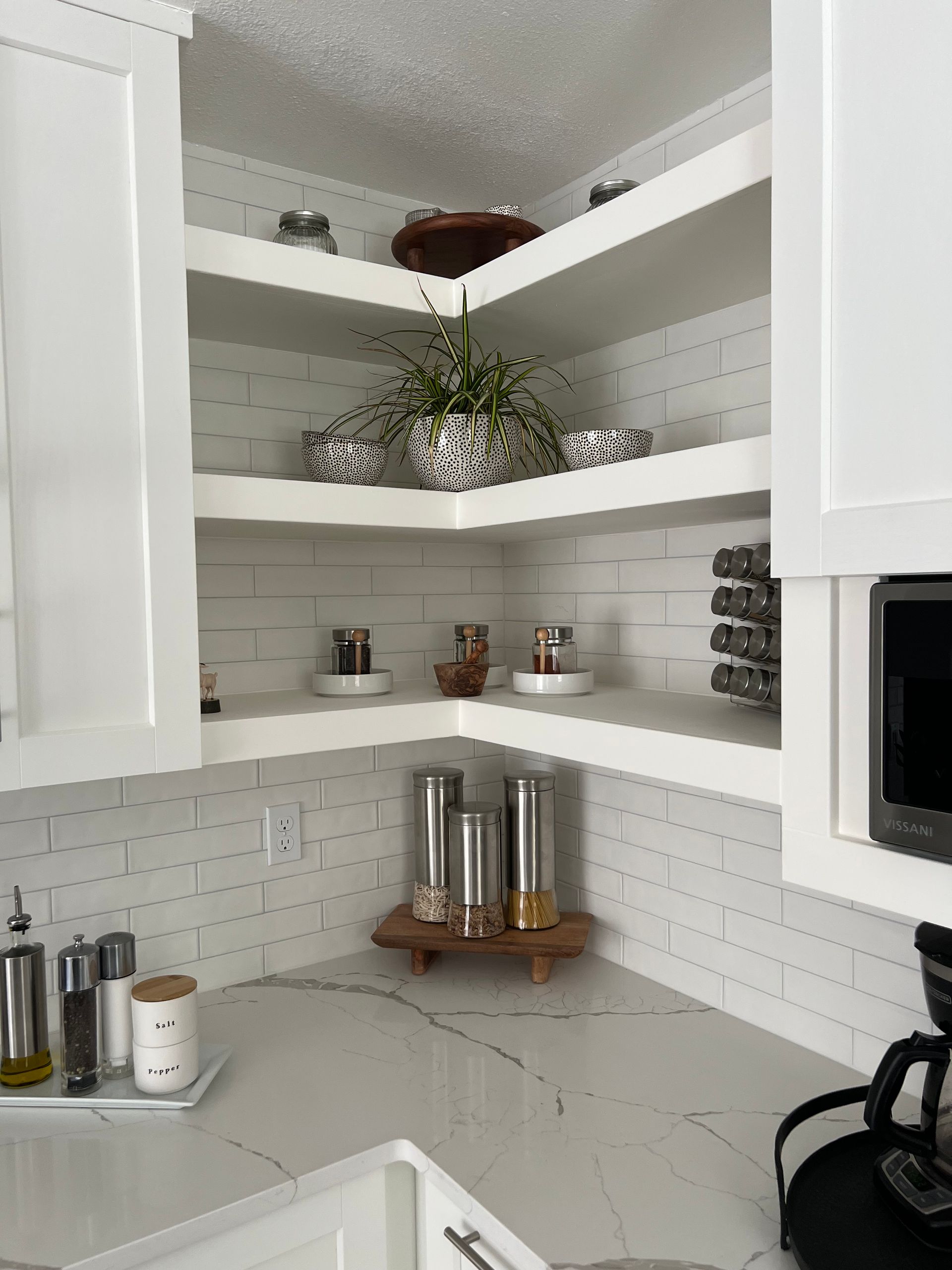 White floating corner shelves hold potted plants and kitchen containers against a white brick backsplash in a kitchen.