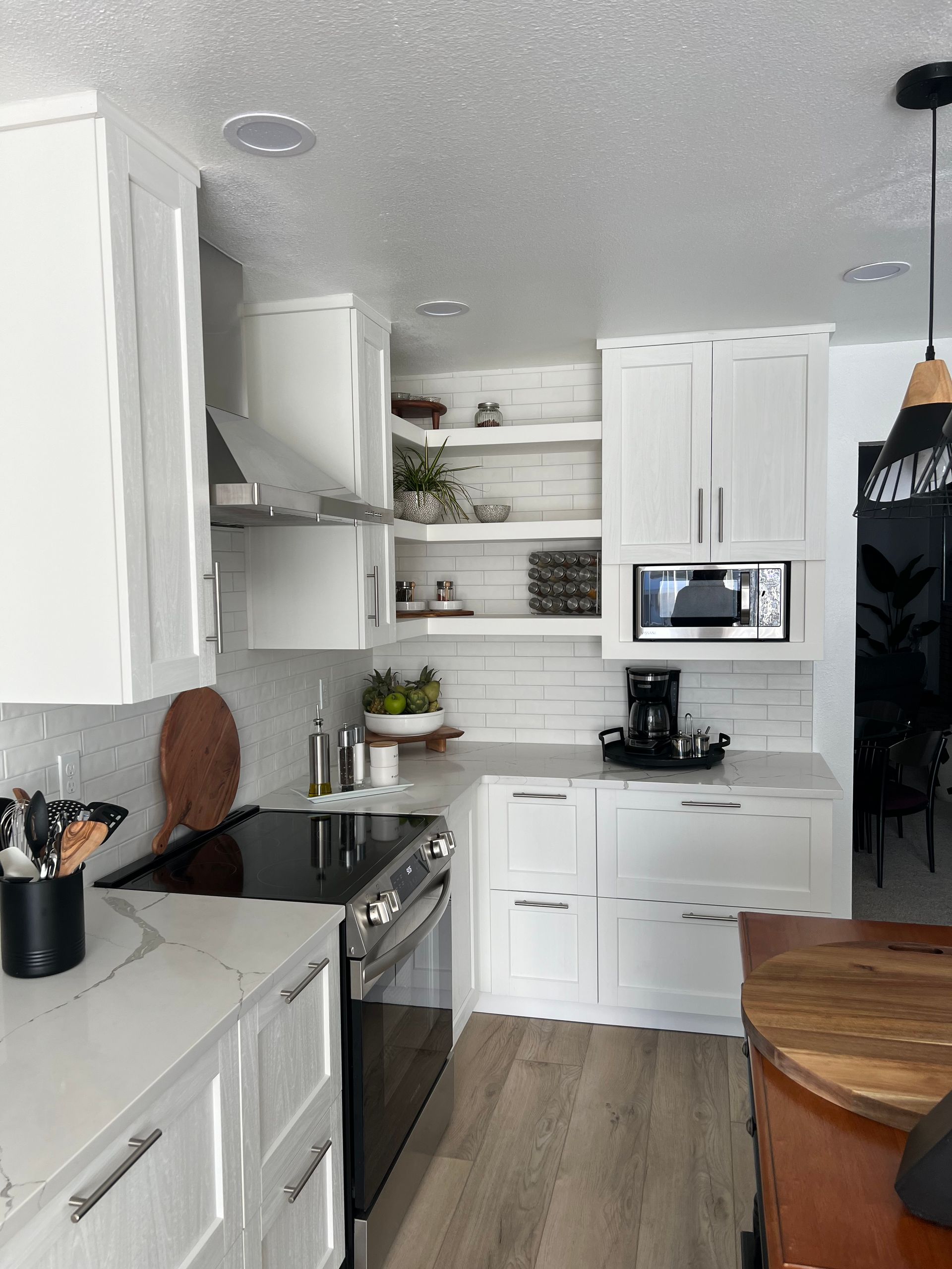 A bright white kitchen with open shelving, a stainless stove, marble countertops, and wood-look flooring.