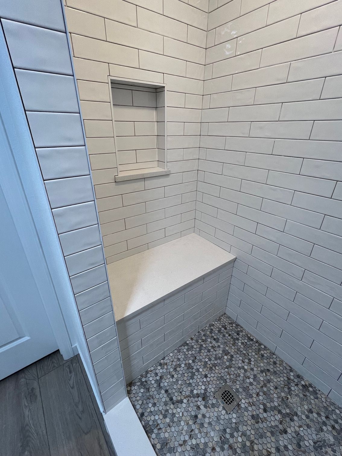 A shower featuring white subway tile walls, a built-in white bench, a recessed niche, and a grey pebble-stone floor.