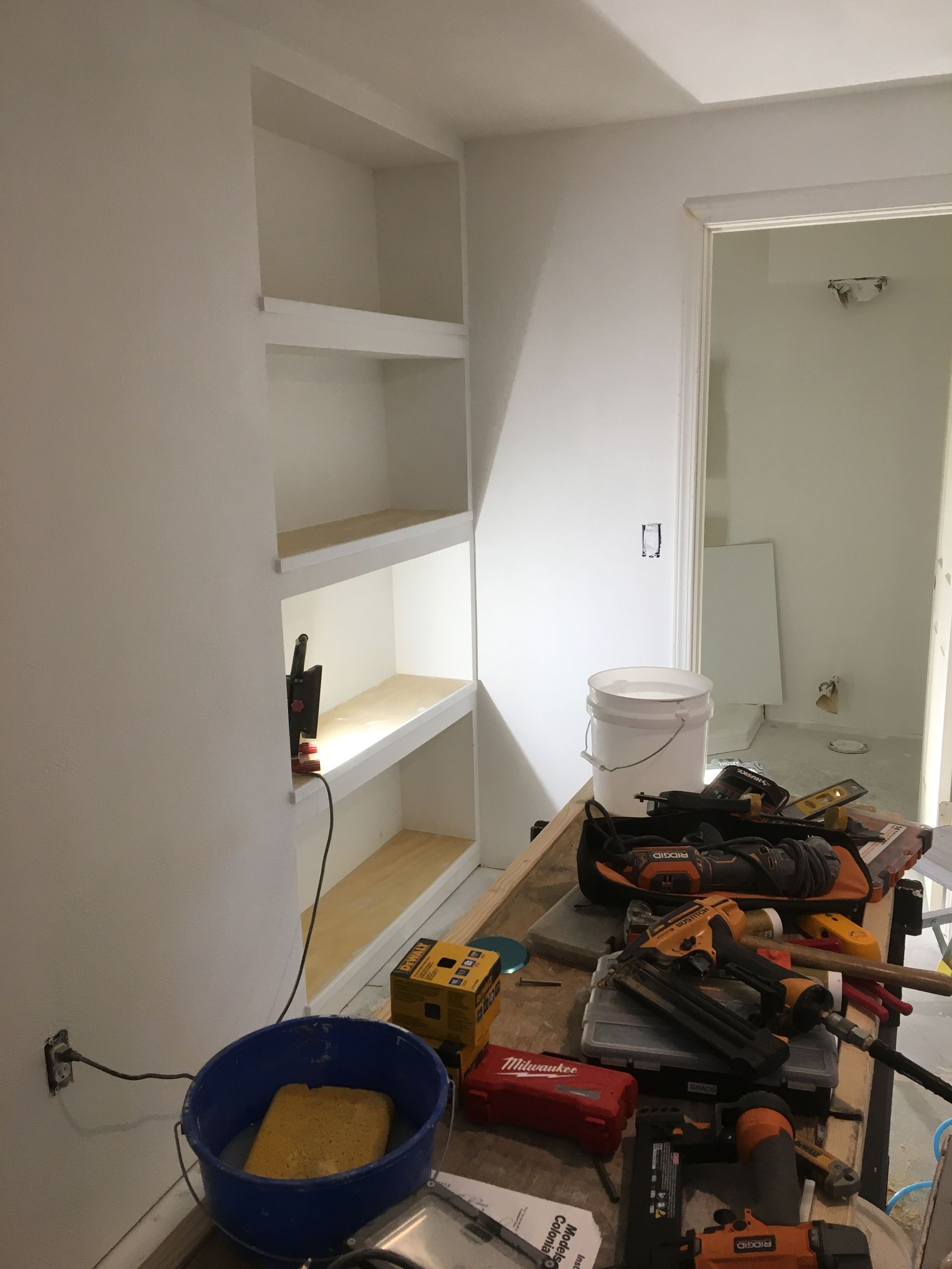 A workspace during construction showing built-in wall shelving and a table cluttered with various power tools.