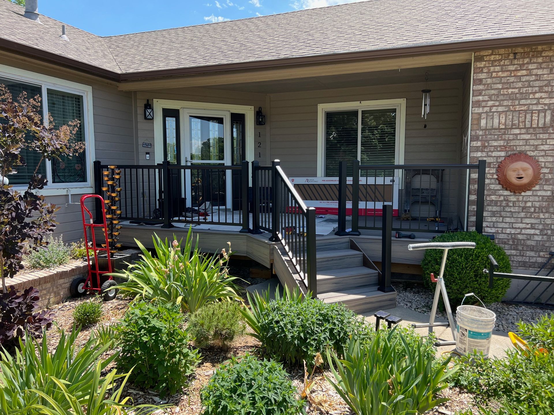 A house exterior featuring a wooden deck with black railings, a red hand truck, and a landscaped garden in the front yard.