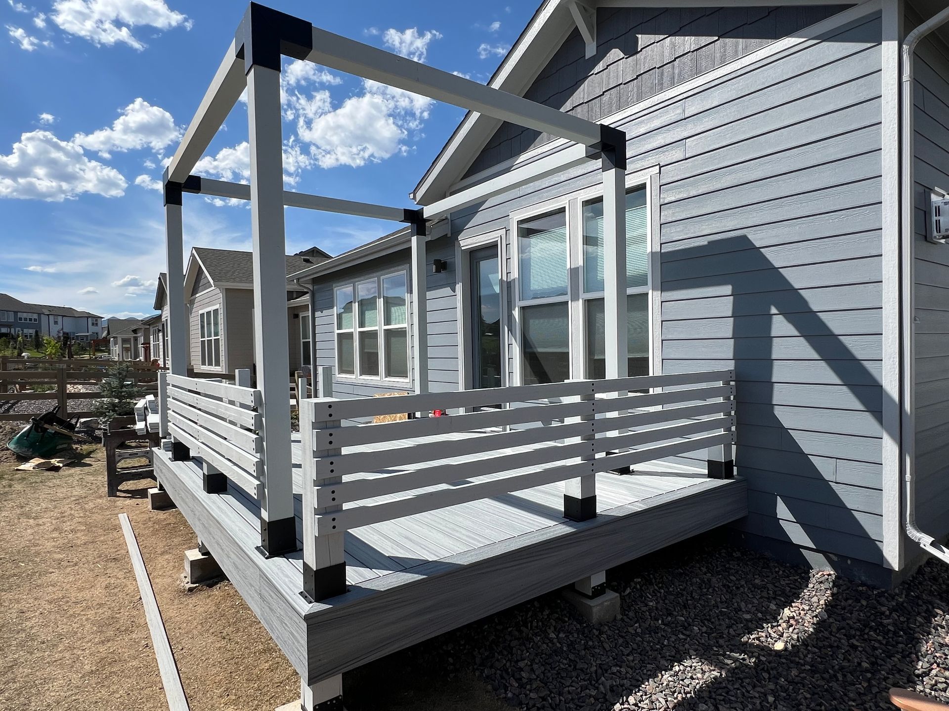 A gray wood deck with a horizontal-slat railing and a modern pergola frame attached to a house under a blue sky.