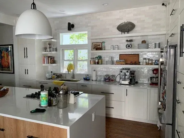 A modern, bright kitchen featuring a white island, wooden base cabinets, open shelving, and a large pendant light.