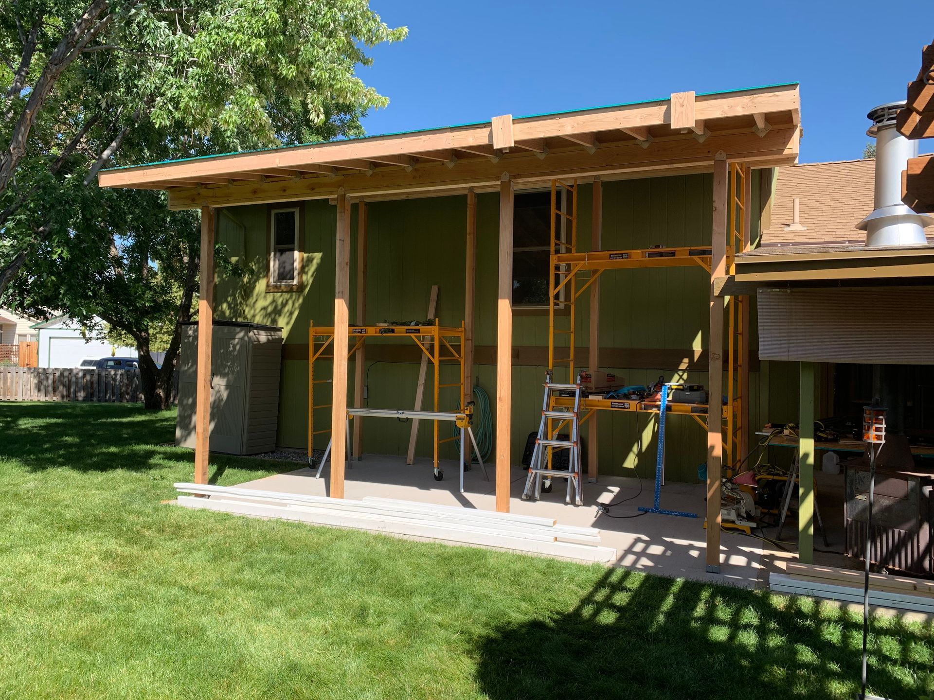 A wooden patio cover structure under construction against the green exterior wall of a house on a sunny day.