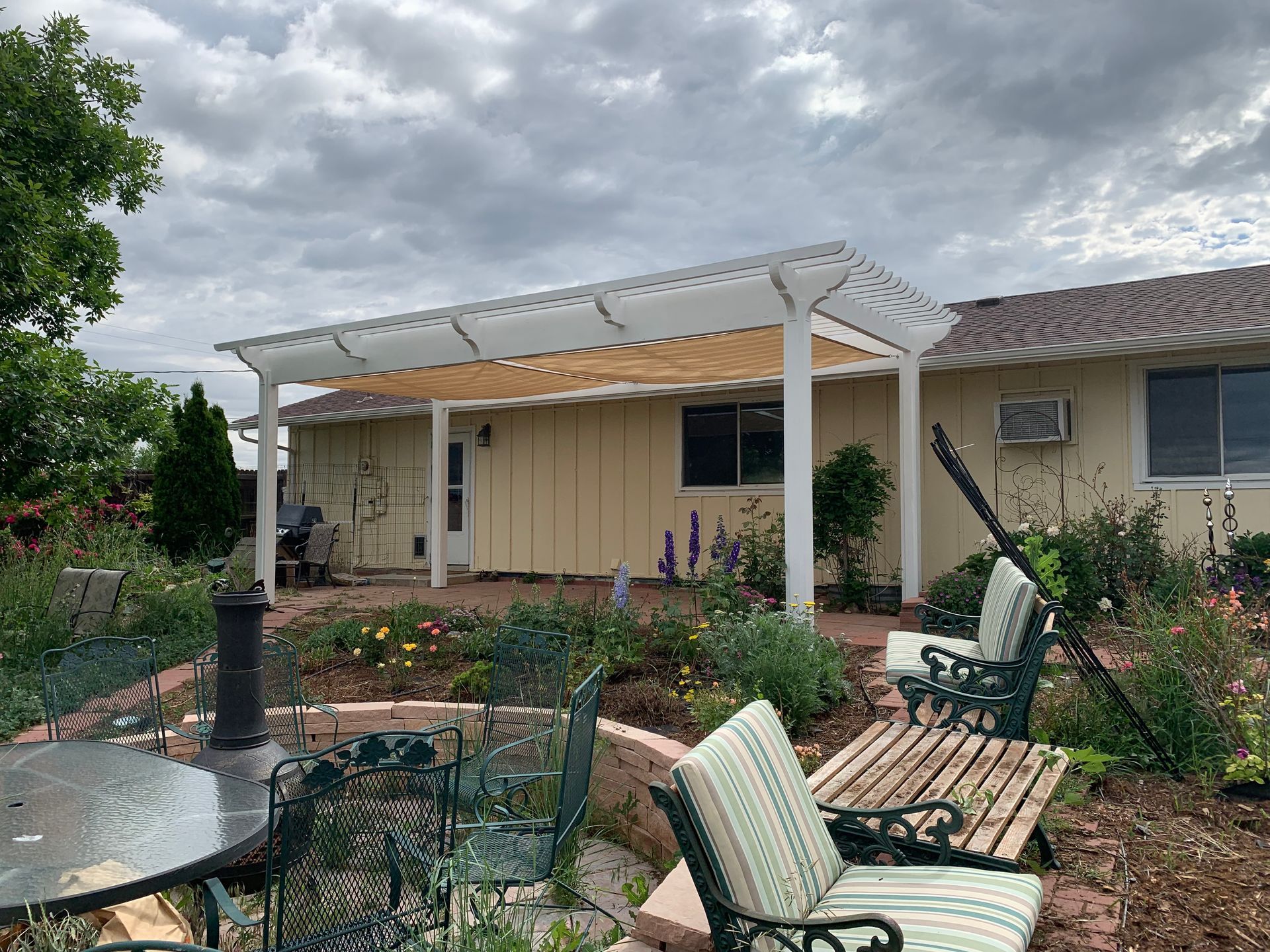 A white pergola with a retractable shade canopy over a patio with outdoor seating in a garden setting.