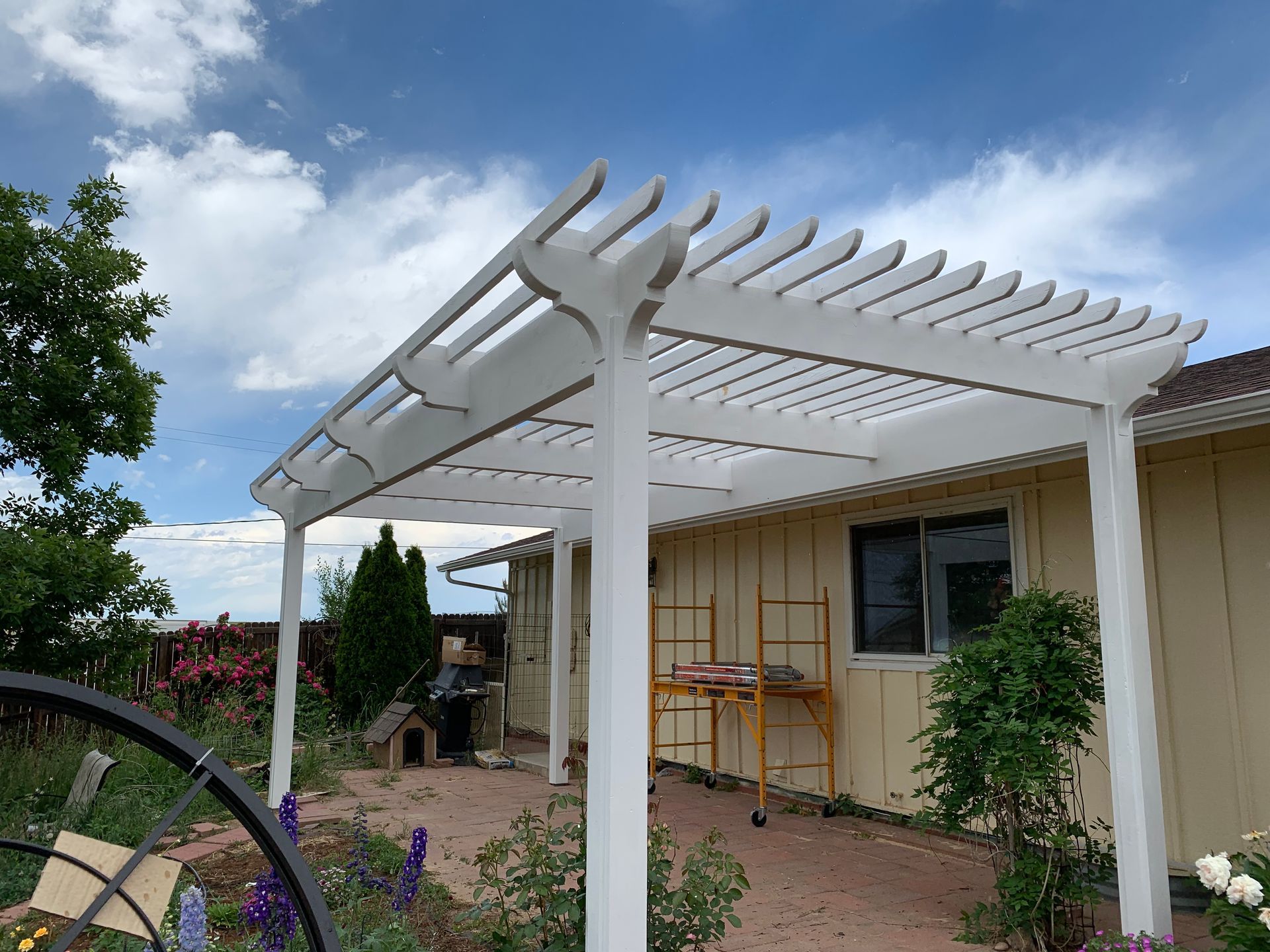 A white wooden pergola stands on a brick patio attached to the side of a yellow house under a blue, cloudy sky.