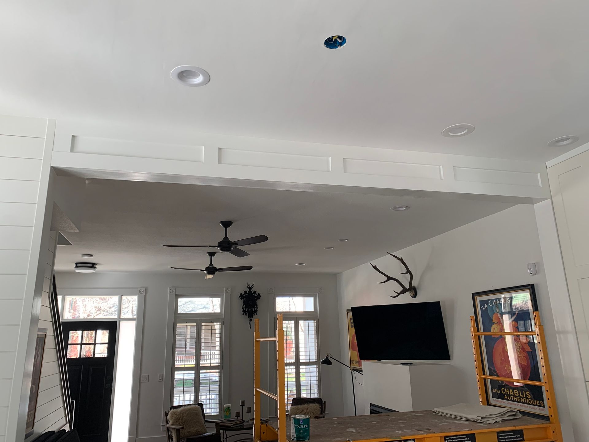 A renovated room with white shiplap walls, recessed ceiling lights, two ceiling fans, and a large television on a stand.