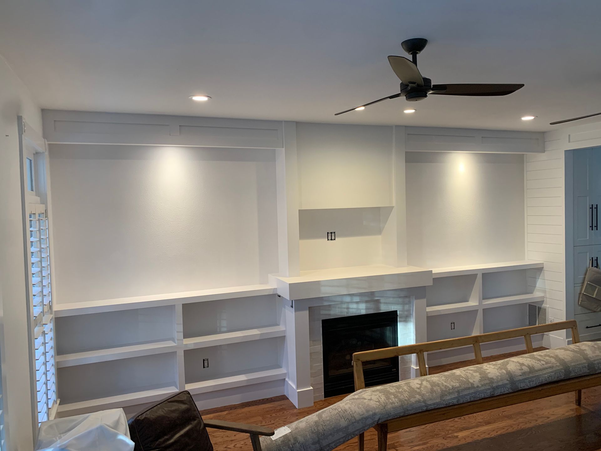 White custom built-in shelves surround a fireplace with a mounted television area and a ceiling fan overhead.