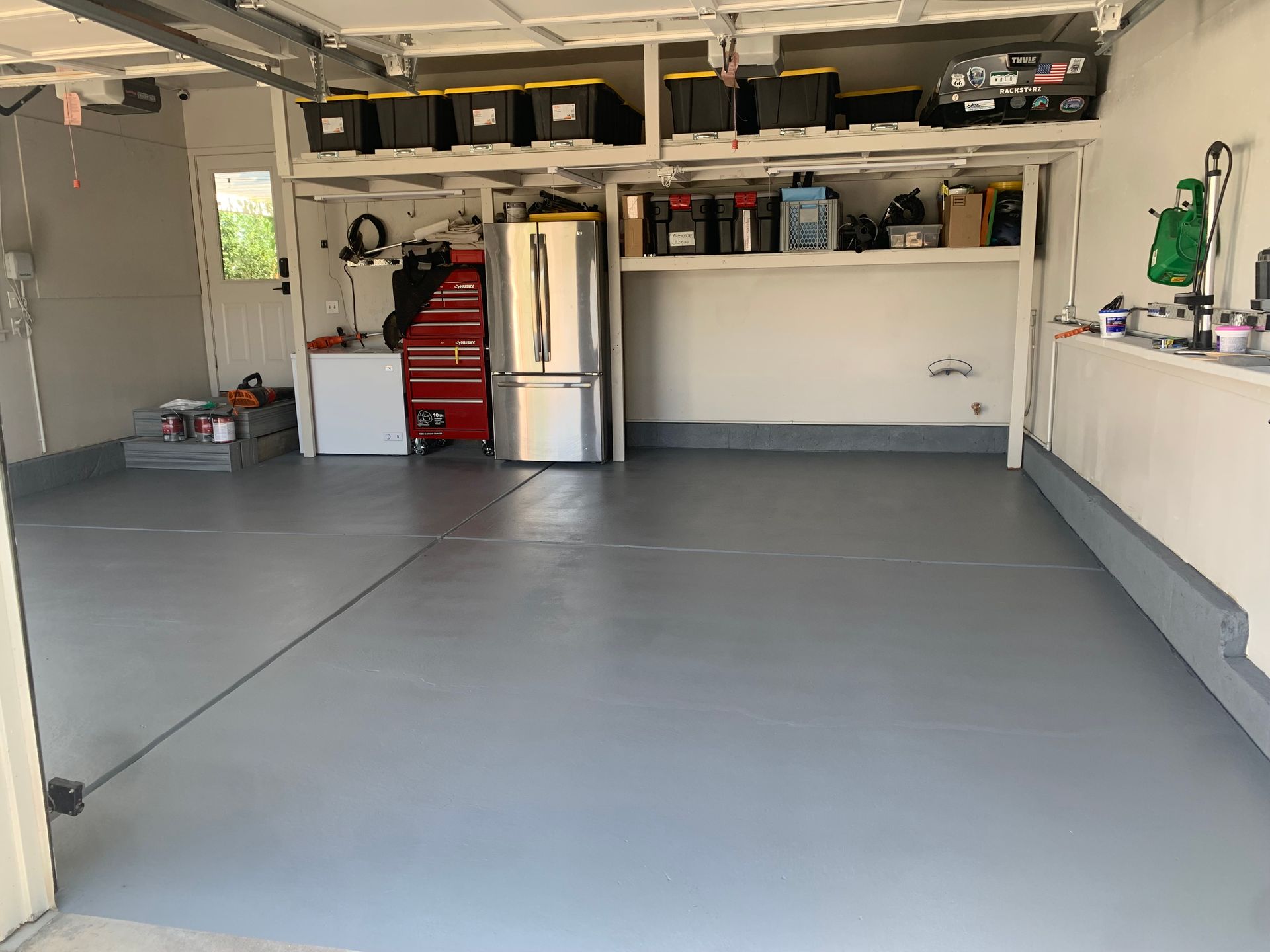 Clean, empty residential garage with gray painted floors, overhead storage shelving, a refrigerator, and a toolbox.