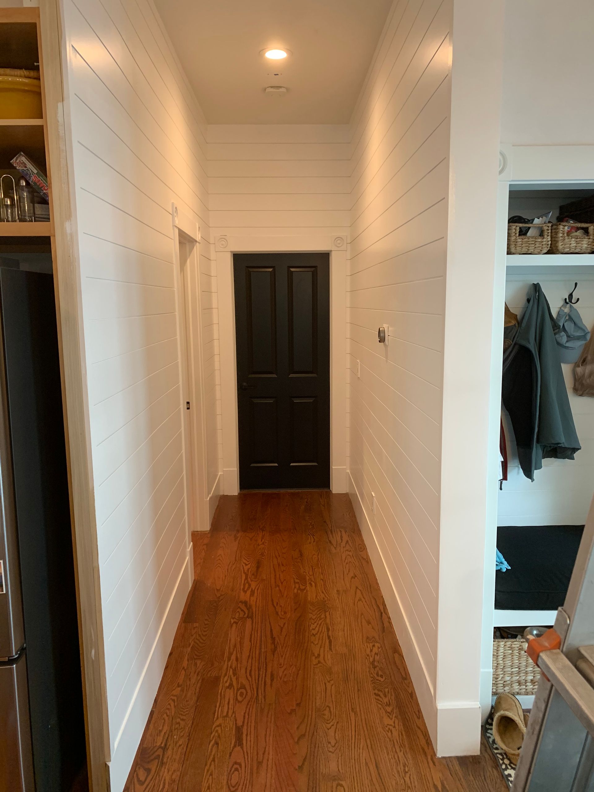 A hallway with white shiplap walls, hardwood floors, and a single black door at the end, next to an open coat storage area.
