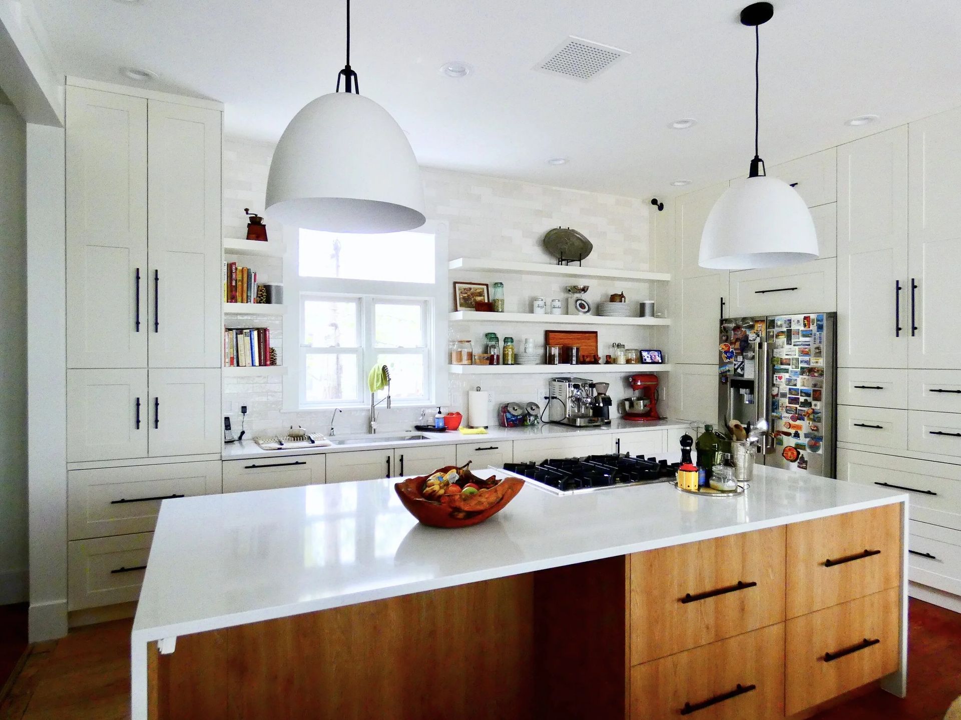 A bright kitchen with white cabinets, light wood island drawers, two large pendant lights, and open shelving.