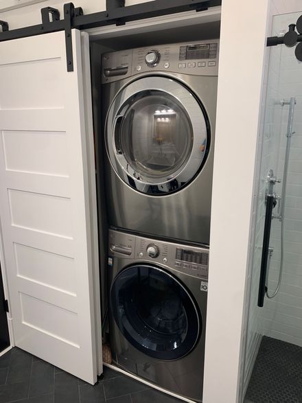 A stacked stainless steel washer and dryer unit installed in a narrow closet with a white sliding barn door.