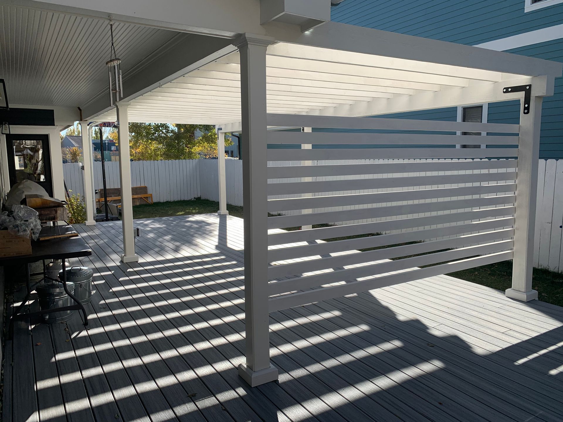 A white pergola with horizontal privacy slats on a gray deck, partially shaded by the structure and a roof overhang.