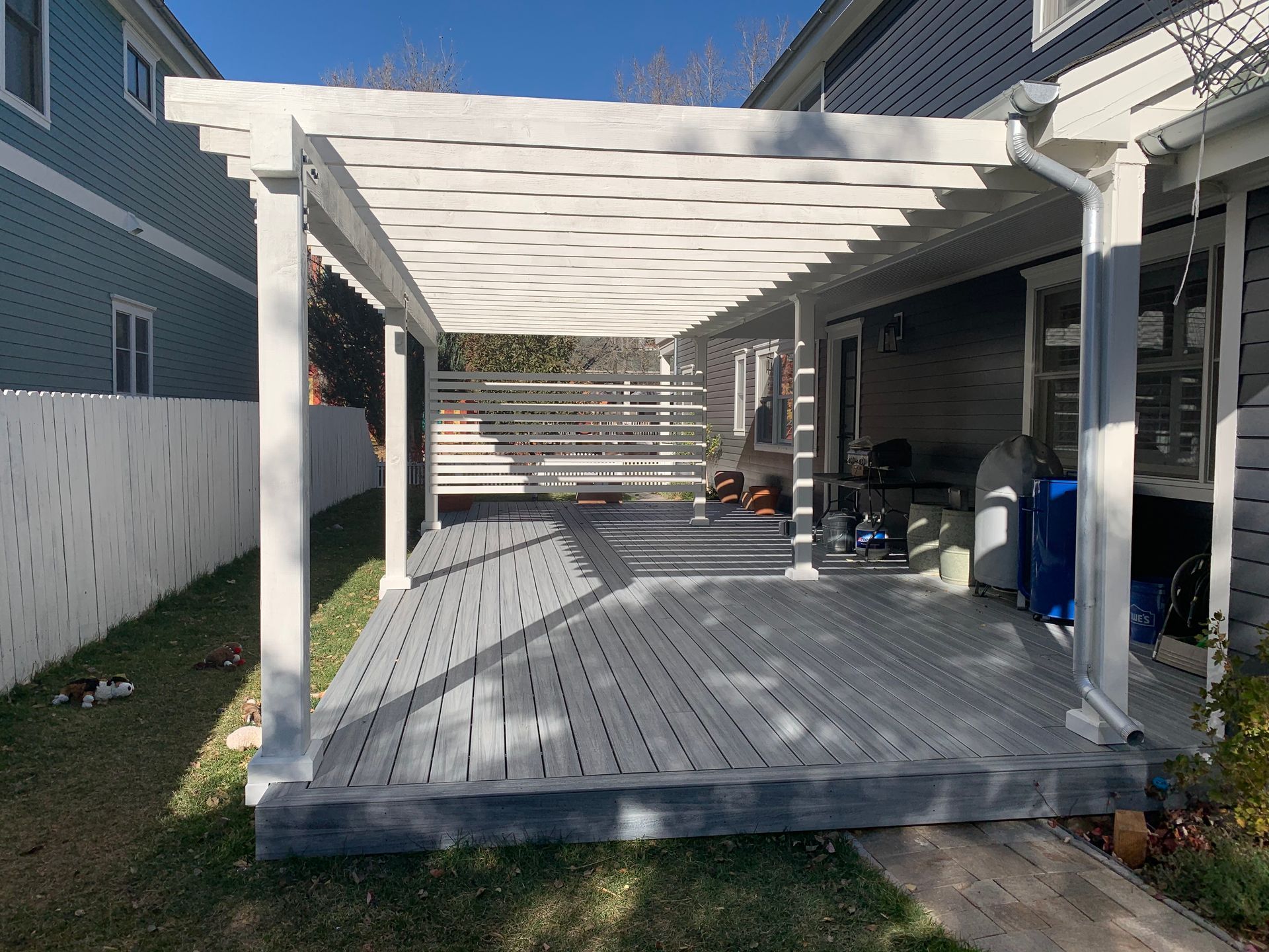 A light-gray wooden deck features a large white pergola extending from a house, with a white fence along the side.