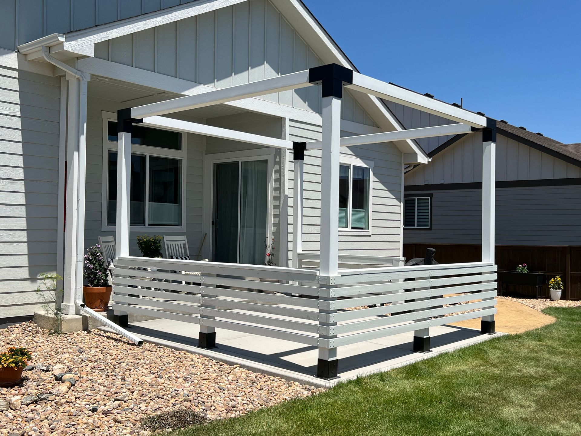 A white pergola with black corner brackets stands on a concrete patio next to a house with white horizontal siding.