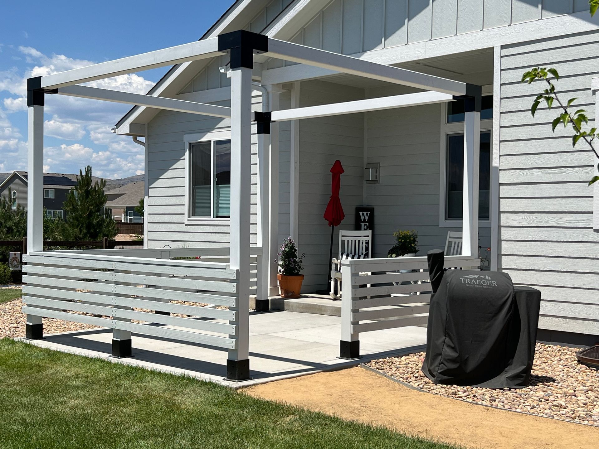 A white pergola with black corner brackets stands on a backyard patio next to a light gray house.