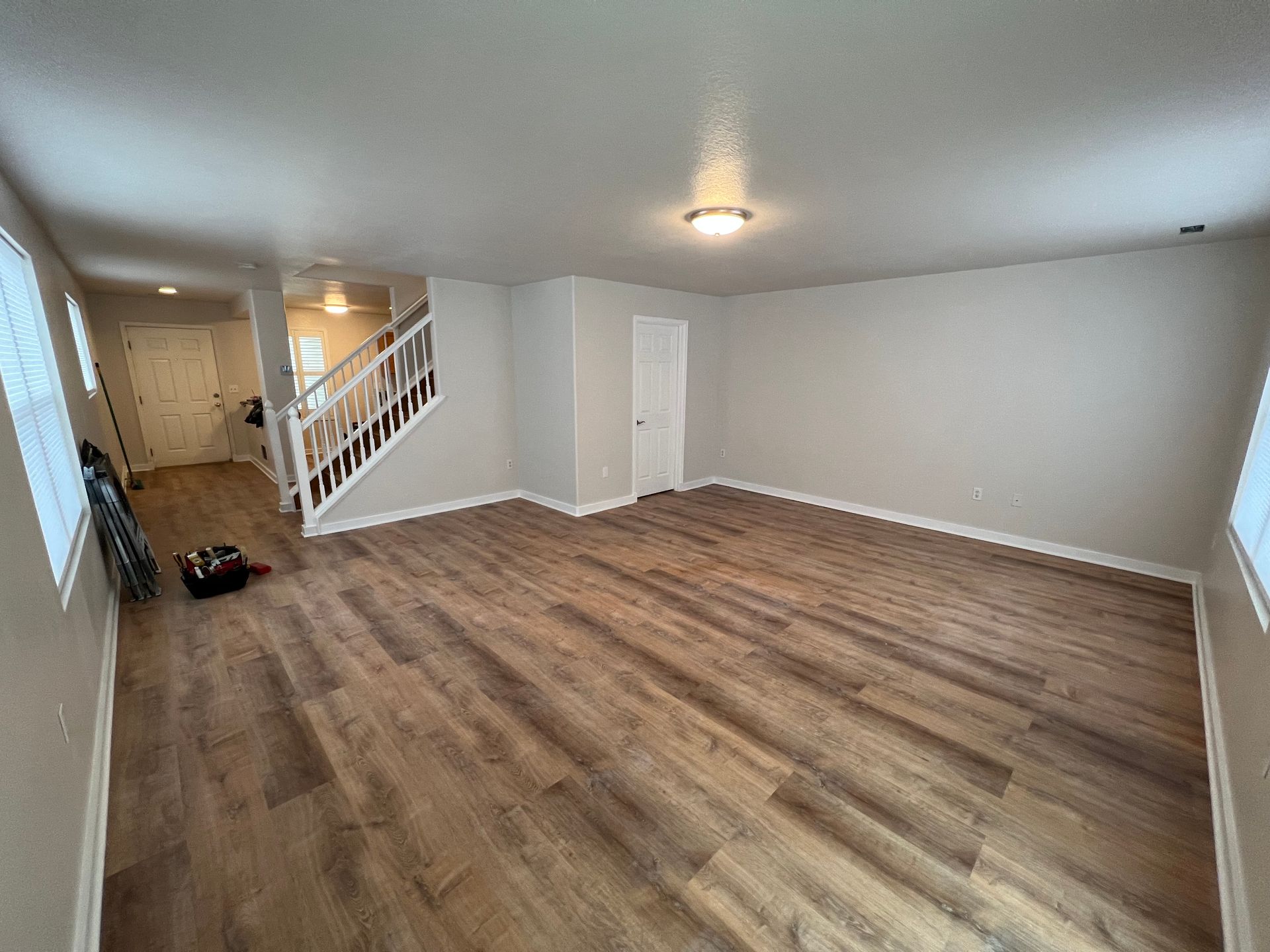 An empty room with light wood-grain vinyl floors, neutral gray walls, a staircase on the left, and a white interior door.