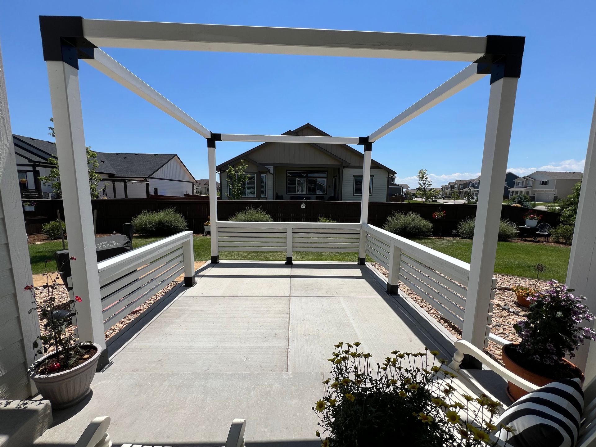 A white pergola frame over a concrete patio with low white railings, set in a backyard on a sunny day.