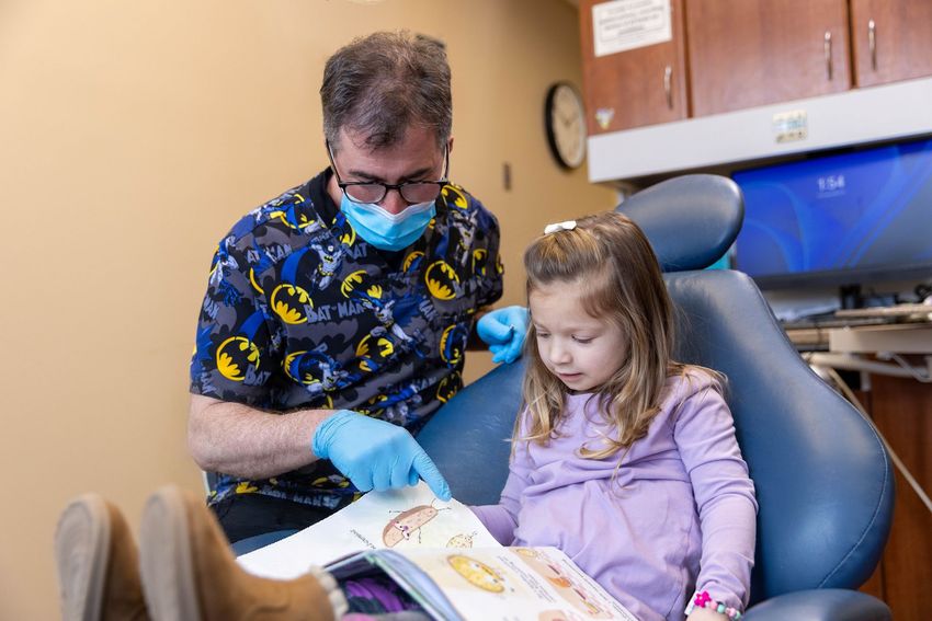 Boy laughing, sitting in a dentist's chair, holding glasses. Turquoise chair, black sweater.