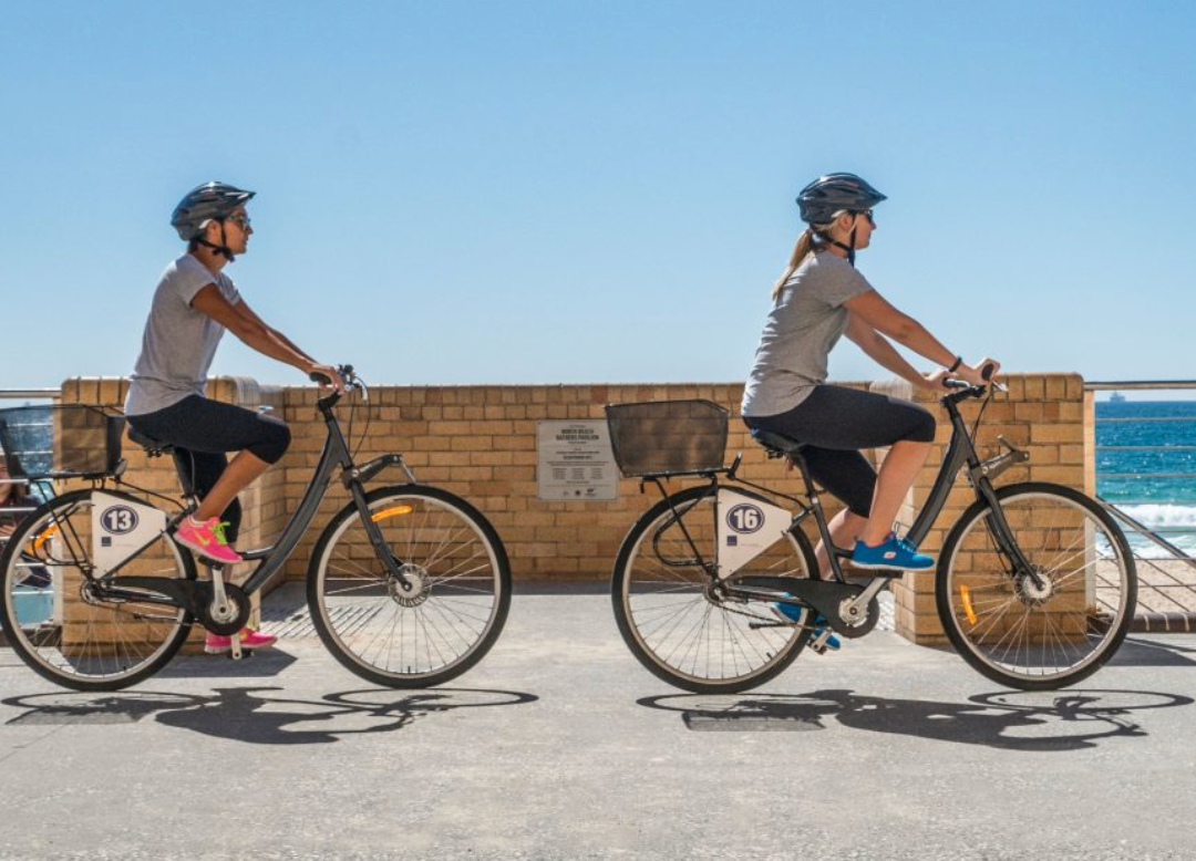 Two women are riding bicycles on a sidewalk near the ocean.