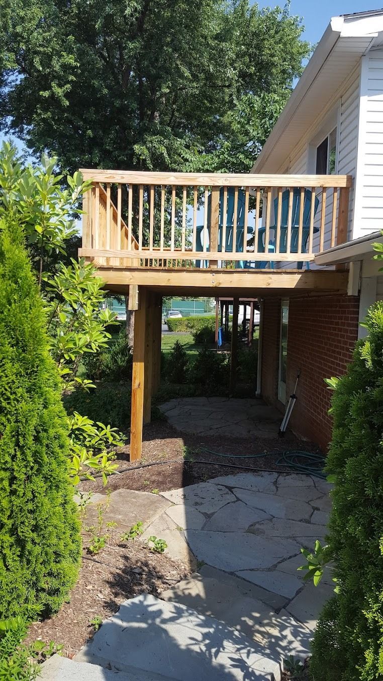 Backyard patio with wooden deck above a stone path, beside a brick house and leafy tree