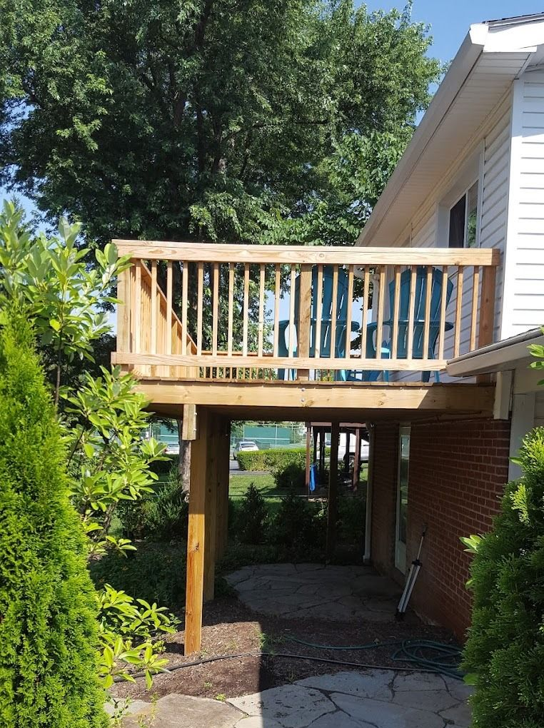 Wooden deck balcony above a driveway beside a house, with trees and shrubs around it.