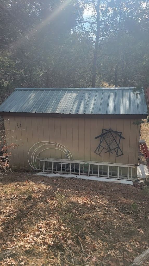 Small beige shed with metal roof in a wooded area, with stacked white chairs and wall art outside.