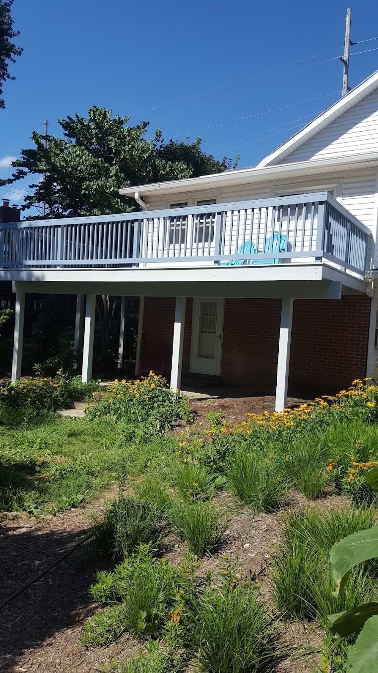 White house with an upstairs deck and white railing above a brick lower level, surrounded by greenery.