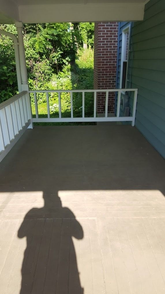 Covered porch with white railing, green siding, and sunlight casting a person’s shadow on the floor