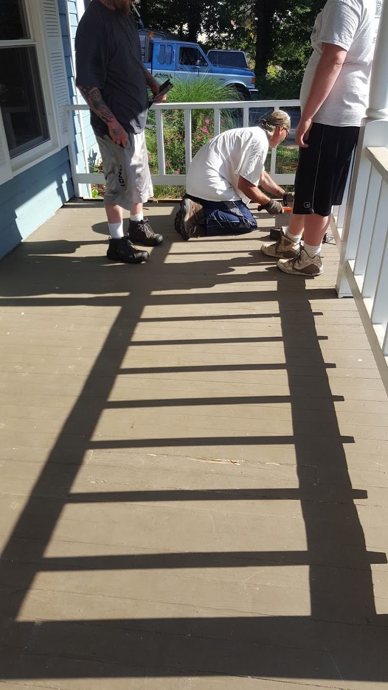 Three people on a porch working near the railing, with a blue car in the background.