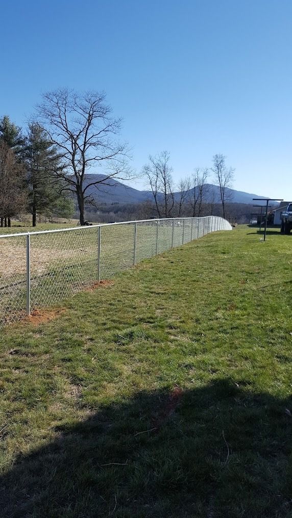 Grassy field beside a chain-link fence, with bare trees, mountains, and a blue sky in the distance.