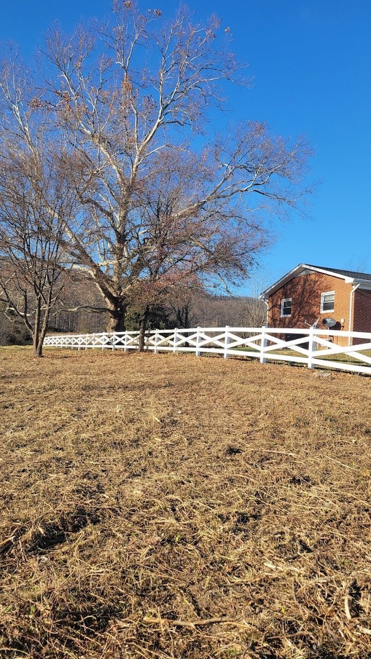 Leaf-covered field with a bare tree, white fence, and brick building under a clear blue sky