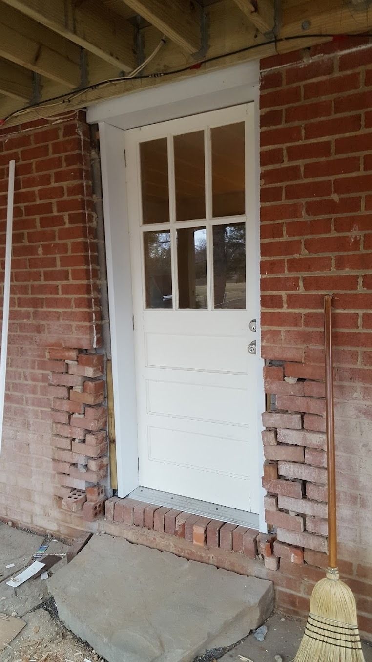 White door set in a red brick wall beneath a porch, with concrete steps leading up to it.