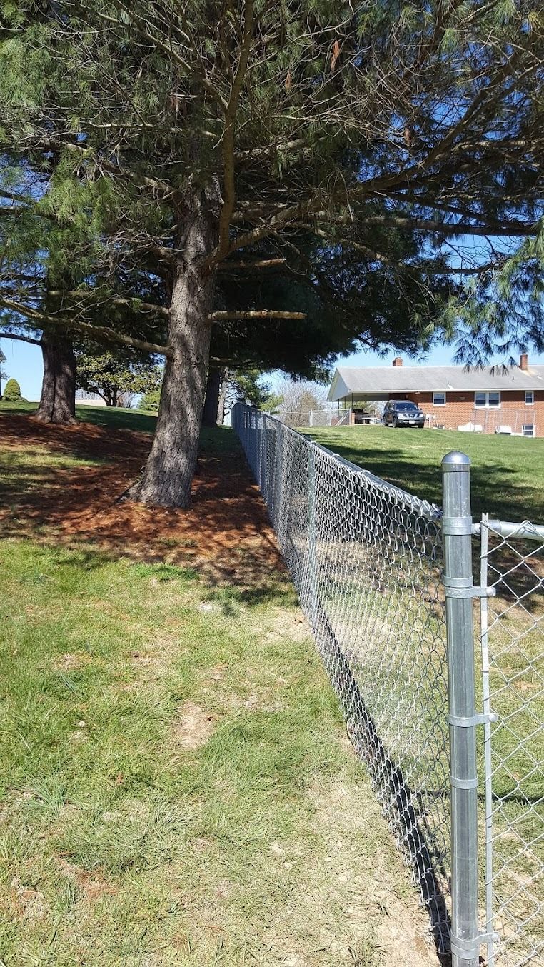 Chain-link fence beside a grassy yard with large trees and a brick building in the background